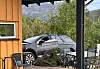 Grå bil parkert ved ei terrasse med møblar og grøne tre.