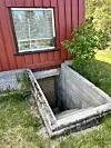 Open kjellaropning ved raud husvegg og gras.