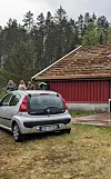 Sølvgrå bil står framfor ei raud hytte ved skogkanten.