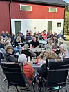 Mange sit rundt bord i ein hage ved ei raud løe.