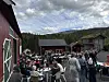 Folk står og sit ute mellom raude bygningar og bord på eit tun.