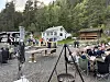 Folk sit ved bord på ei gruslagt uteplass ved eit kvitt hus i skogen.