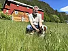 Mann kneisar med hund i høgt gras framfor raud gard.