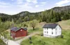 Raudt og kvitt hus på grønt jorde med skog og ås i bakgrunnen.