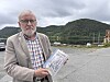 Mann held ei bok ved ein parkeringsplass med fjell i bakgrunnen.
