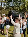 Barn og vaksne peikar oppover ved ei hytte i ein park.