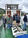 Sju personar står på eit båtdekk med kabin, med vatn og fjell i bakgrunnen.