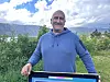 Mann i blå genser står ved eit skilt med fjell og busker i bakgrunnen.