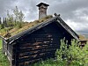 Svartna tømmerhus med torvtak i grønt landskap.
