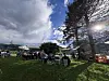 Folk sit på stolar og ved bord på ein plen med tre og sol bak.