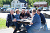 Seks personar sit rundt eit piknikbord ute ved eit kulturhus og snakkar saman.