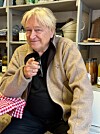 Mann sitt ved bord og peikar mot kamera i ein butikk.