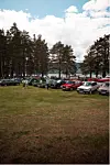 Bilar står parkert på ei grasmark ved skog og vatn.
