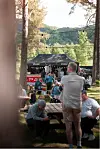 Folk sit og står ved bord og benker ved ein utandørs bar i ein park.