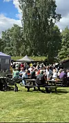Folk sit ved bord og benkar under telt i ein park.