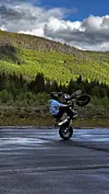 Motorsyklist på bakhjulet på våt veg med skog og fjell bak.