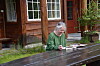 Kvinne i grønn genser sit ved utebord framfor trehus.