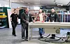 Fire menn står rundt ein arbeidsbenk i eit industrilokale.