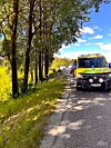 Gul ambulanse står ved vegkant med skog på venstre side og bilar lenger framme.