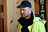 Mann i gul refleksjakke og svart caps står ved ei open dør.