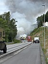 Brannbil og utrykkingskøyretøy ved røykfylt veg i kupert landskap.