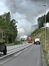 Brannbil og utrykkingskøyretøy ved røykfylt veg i kupert landskap.