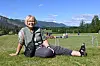 Ein person sit på graset ved ei fotballbane med fjell i bakgrunnen.