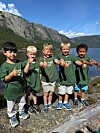 Fem barn står på ein steinmur ved ei innsjø og viser tommelen opp.