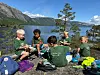 Fem barn sit på steinar ved eit vatn og les i bøker.
