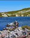 Vrakrestar ligg i steinrøys ved blått fjordvatn.