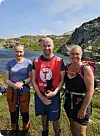 Tre personar står med turutstyr ved sjøen i solskin.