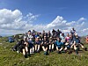 Stor gruppe personar på ei grasdekt fjelltopp under blå himmel.