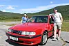 To personar står ved ein raud bil på ein vegplass med fjell i bakgrunnen.