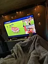 TV-skjerm med fotballbilete i eit mørkt rom, sett over ein sofa med tepper.