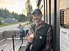 Mann med drikke står utanfor ei kro, med to personar på terrassen bak.