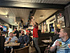 Gjestar sit i ein bar medan ein mann i raud genser står med armane i vêret.