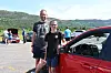 To personar står ved ein raud bil på ein parkeringsplass med fleire bilar og fjell bak.