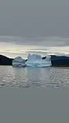 Stort isfjell flyt i vatn framfor fjell og skya himmel.