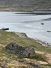 Lite seglfartøy på stille vatn ved grasdekt strand og steinruin.