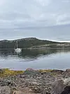 Seglbåt på stille vatn ved steinstrand og skogkledd kyst.