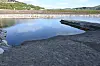 Stille fjordbasseng med steinstrand, molo og grøne åsar.