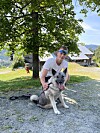 Mann med solbriller sit på bakken og held ein stor hund framfor eit gardshus.