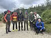 Sju turfolk og ein svart hund står samla på ein grusveg ved skog og fjell.