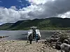 To personar står framfor ein båt ved ei steinstrand med fjell og skyer.