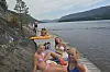 Fire personar kviler på ei steinstrand ved vatnet, med kajakkar og fjell bak.