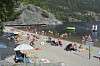 Folk oppheld seg på ei steinstrand ved vatnet med båtar og fjell i bakgrunnen.