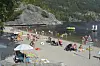 Folk oppheld seg på ei steinstrand ved vatnet med båtar og fjell i bakgrunnen.