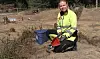 Person på kne ved blå bøtte og raud spade på ein jordete arbeidsplass.