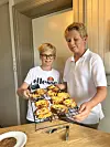 To gutar held eit steikebrett med muffins ved eit bord.