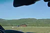 Raud hytte i grasmark med fjell i bakgrunnen, sett frå ein bil.